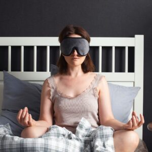 Woman sitting in her bed meditating with a night eye mask on.